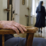 Main âgée sur un fauteuil avec silhouette de religieuse en arrière-plan dans un couloir de maison de retraite.