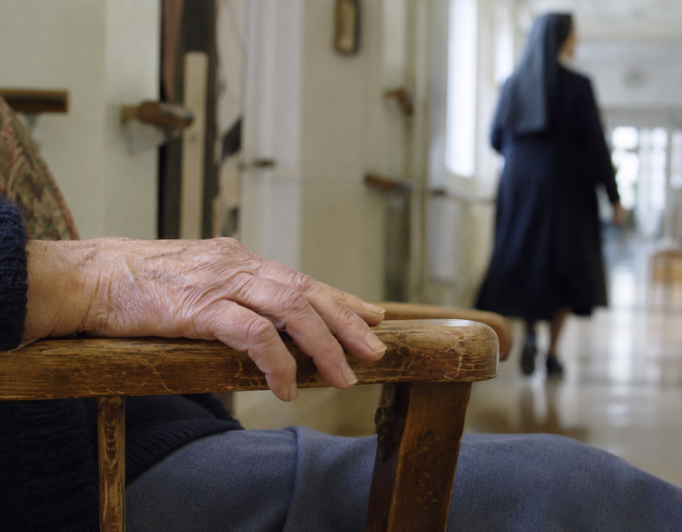 Main âgée sur un fauteuil avec silhouette de religieuse en arrière-plan dans un couloir de maison de retraite.