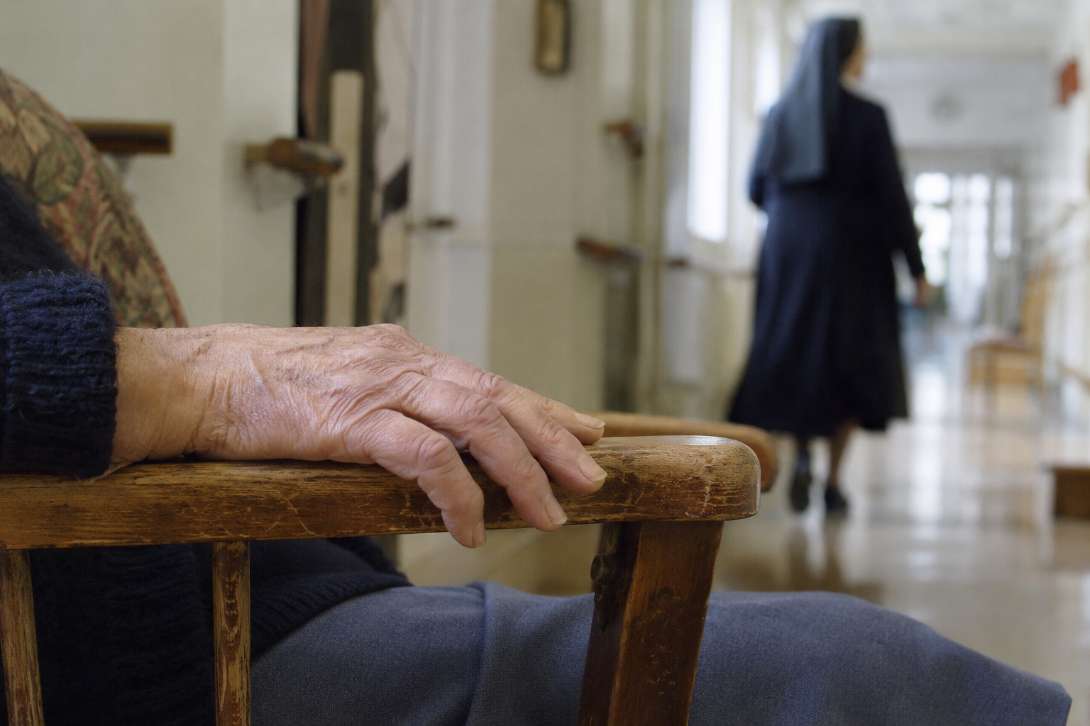 Main âgée sur un fauteuil avec silhouette de religieuse en arrière-plan dans un couloir de maison de retraite.