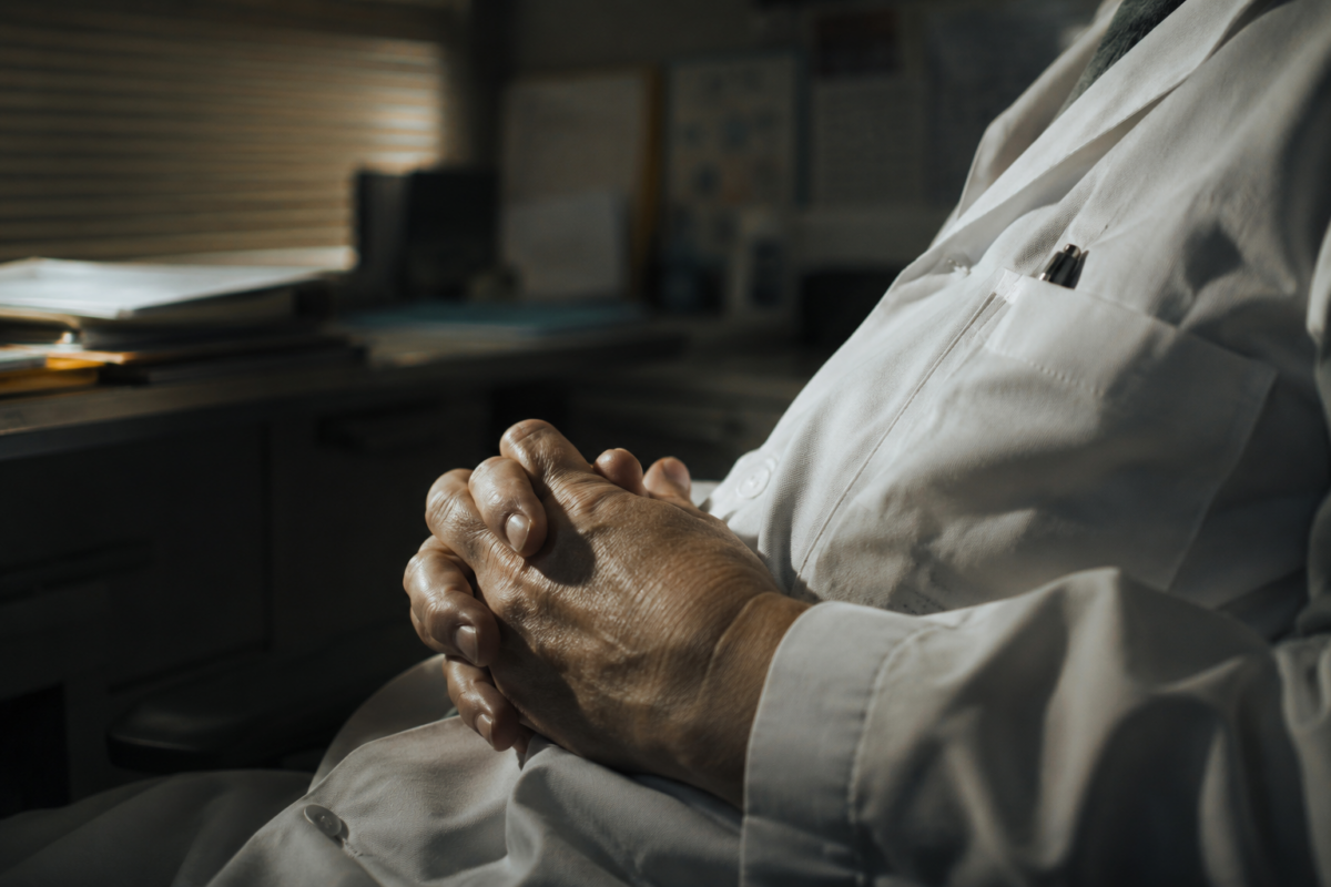 Les mains croisées d'un médecin âgé portant une blouse blanche dans un bureau sombre.