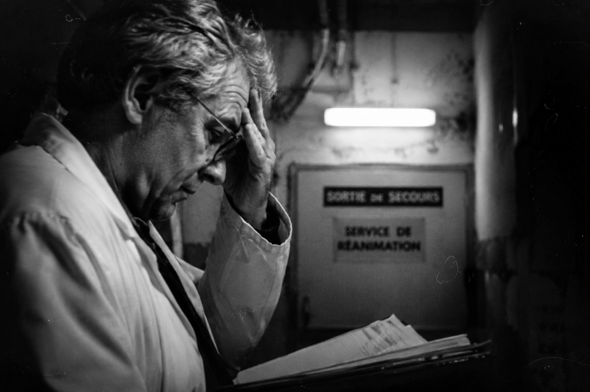 Médecin fatigué en blouse blanche tenant un dossier médical dans un couloir d'hôpital sombre, symbolisant la liberté de conscience face à l'aide à mourir.