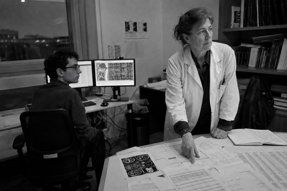 Une médecin examine un dossier tandis qu’un chercheur analyse des données médicales sur deux écrans à proximité.
