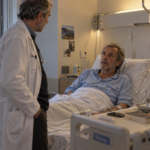 Un patient en pyjama et perfusé écoute son médecin debout au bord de son lit.